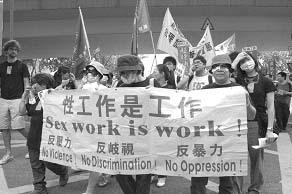 A black and white photo depicts several people carrying a fabric banner in front of them, and others following behind. Several people are wearing masks to cover part of their faces. On the banner, the English text is "Sex work is work! No violence! No discrimination! No oppression!" There's also some writing not in English - presumably in whatever alphabet or system is common in Hong Kong.