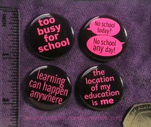 Photo of four badges. 1. "too busy for school". 2. "No school today?" "No school any day!" with each phrase shown on a speech bubble. 3. "learning can happen anywhere". 4. "the location of my education is me". On each badge, the design is pink on a black background. A ruler is shown nearby, and each badge is about 1 inch in diameter. In the corner is the edge of a 50p coin. The background is a piece of purple velour.