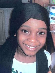 Photo of Khyra Ishaq. A young Black girl is smiling at the camera. She has baby teeth and brown eyes. She is wearing a black headscarf. She looks happy.