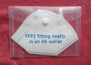 FFP2 fitting neatly in an A6 wallet. The mask is seen from the side, folded flat. It's in a transparent plastic wallet which has a popper to hold it closed.