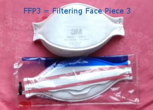 FFP3 = Filtering Face Piece 3. A shaped white mask, seen from the front. Another similar one seen from the back, still in its plastic packet, showing two rubber bands which are designed to go behind the head.