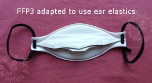 FFP3 adapted to use ear elastics. A shaped white mask seen from the angle where you'd be about to put it on, with a narrow grey foam strip across the nose area. It has black elastic ear loops.
