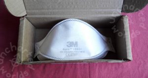 An FFP3 respirator mask tucked into a small cardboard box. The mask is in an unflattened three-dimensional shape ready to be put on. It's white with a 3M logo in faint grey on the front, a CE mark "CE 2797" and a few other numbers..